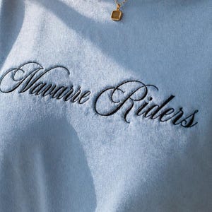 Puede incluir: Una camiseta azul claro con el texto bordado "Navarre Riders" en escritura negra. Se lleva un collar de oro con un colgante cuadrado. La camiseta es de material suave.