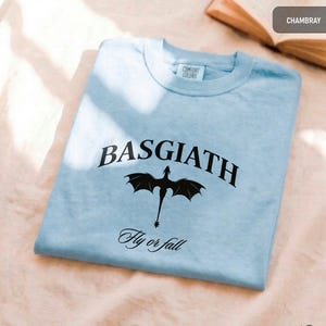 Peut inclure: T-shirt bleu clair avec le mot "BASGIATH" en arc de cercle et un dessin de dragon noir en dessous. L'inscription "Fly or fall" est imprim&eacute;e en dessous. Marque Comfort Colors.