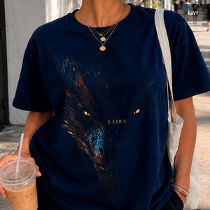 May include: Navy blue t-shirt featuring a detailed graphic of a wolf's face with glowing yellow eyes. The word "TAIRN" is printed below the image. The shirt is short-sleeved and made of a soft material.
