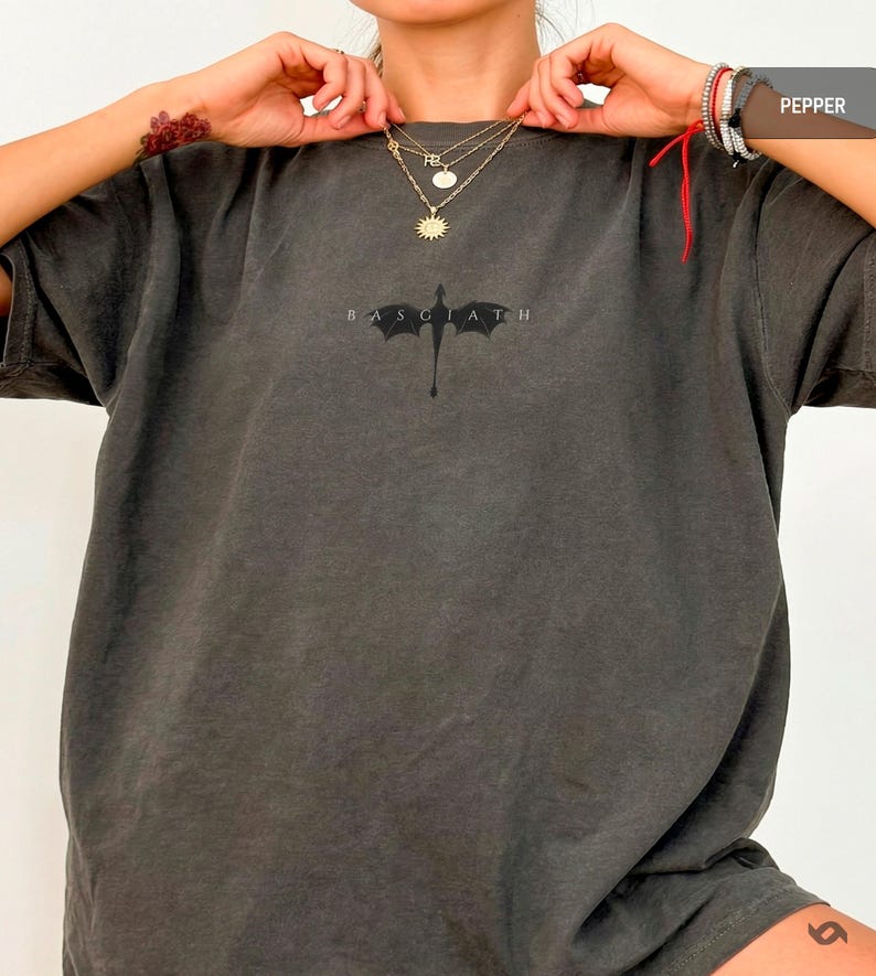 May include: Dark grey t-shirt featuring a dragon graphic and the word "BASGIATH" underneath. The shirt has a faded appearance. The model is wearing gold necklaces.