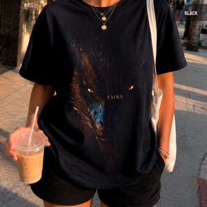 May include: Black t-shirt featuring a detailed graphic of a dragon's face with glowing yellow eyes and the word "TAIRN" in a serif font. The shirt is paired with black shorts and a tote bag.