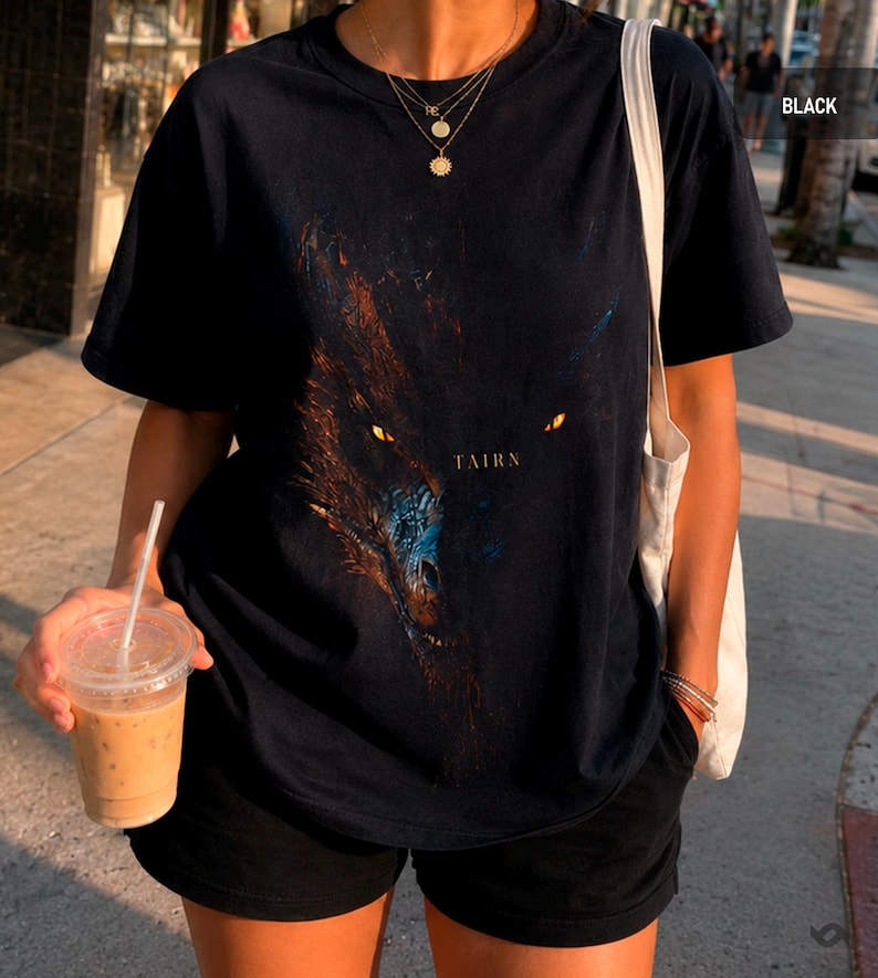 May include: Black t-shirt featuring a detailed graphic of a dragon's face with glowing yellow eyes and the word "TAIRN" in a serif font. The shirt is paired with black shorts and a tote bag.