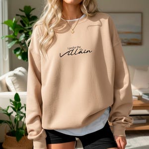 May include: A tan sweatshirt with the text "I prefer the Villain" in black script. The sweatshirt has a crew neck and long sleeves. The model is wearing black shorts and a white undershirt.