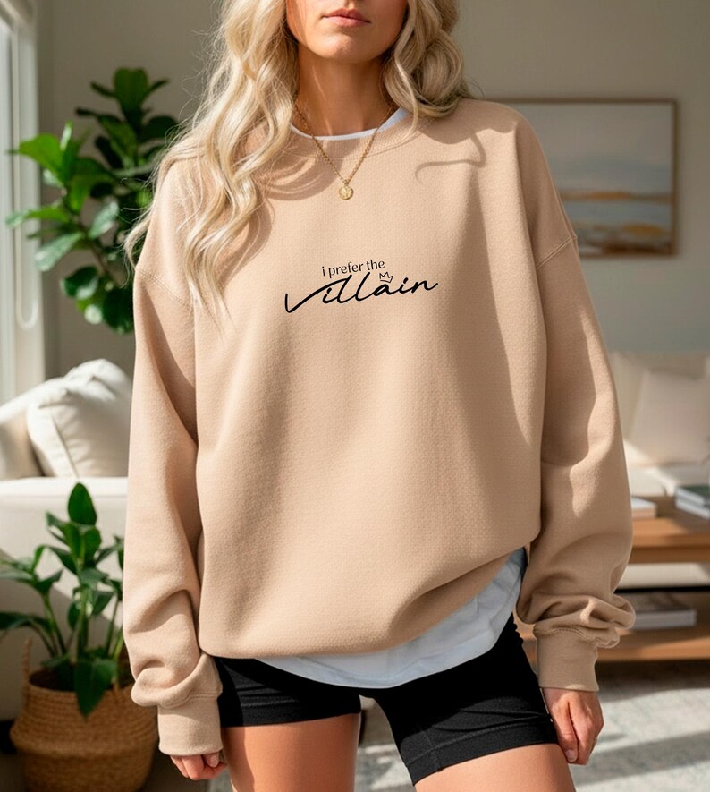 May include: A tan sweatshirt with the text "I prefer the Villain" in black script. The sweatshirt has a crew neck and long sleeves. The model is wearing black shorts and a white undershirt.