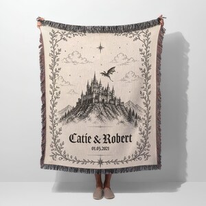 Op de afbeelding: Cr&egrave;mekleurige geweven plaid met een zwart-witte kasteelillustratie, een vliegende draak en de namen "Catie & Robert" met een datum. De plaid heeft een decoratieve rand en franjes.