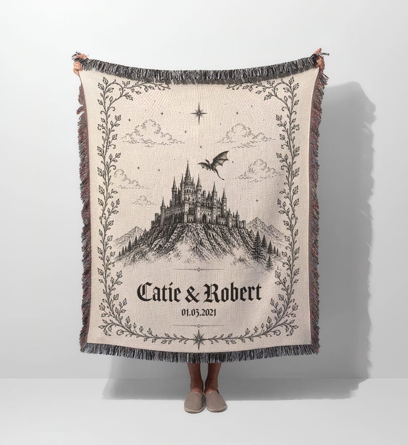Op de afbeelding: Cr&egrave;mekleurige geweven plaid met een zwart-witte kasteelillustratie, een vliegende draak en de namen "Catie & Robert" met een datum. De plaid heeft een decoratieve rand en franjes.