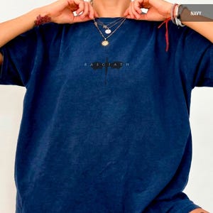 May include: Navy blue t-shirt featuring the text "BASGIATH" and a graphic. The shirt is a solid colour and has a crew neck. The model is wearing layered necklaces.