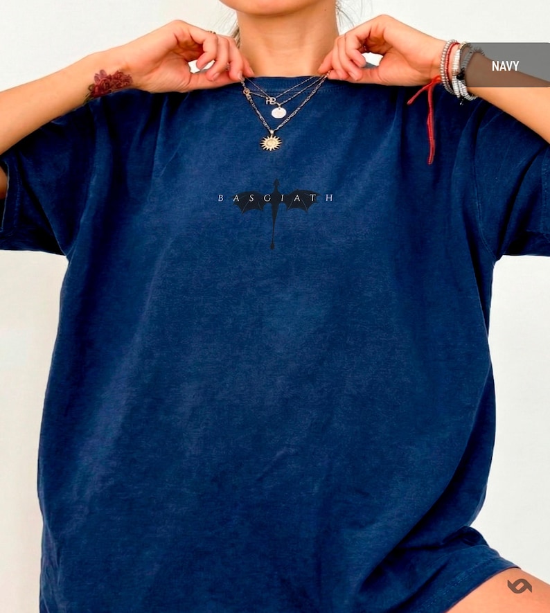 May include: Navy blue t-shirt featuring the text "BASGIATH" and a graphic. The shirt is a solid colour and has a crew neck. The model is wearing layered necklaces.