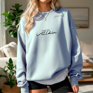May include: A light blue sweatshirt with the text "I prefer the Villain" in black script. The long-sleeved top has a crew neck and is worn with black shorts. The sweatshirt is a solid color and appears to be made of a soft material.