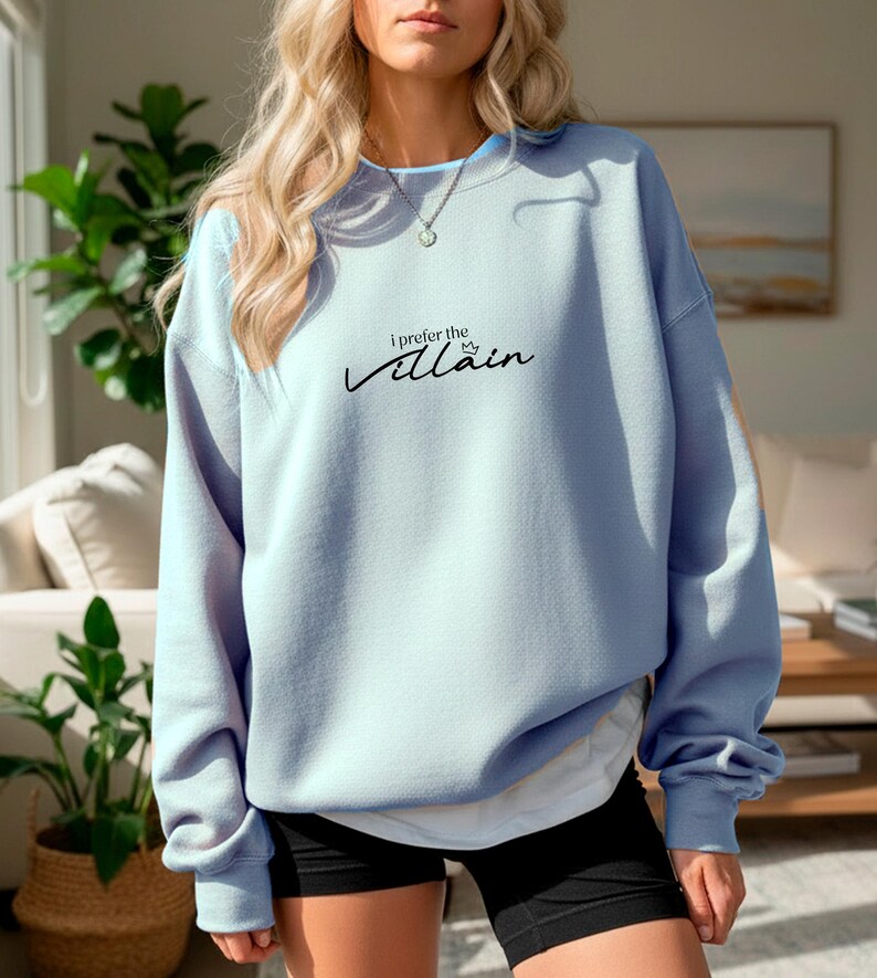 May include: A light blue sweatshirt with the text "I prefer the Villain" in black script. The long-sleeved top has a crew neck and is worn with black shorts. The sweatshirt is a solid color and appears to be made of a soft material.