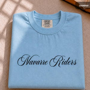Puede incluir: Una camiseta azul claro con las palabras "Navarre Riders" en cursiva. La camiseta est&aacute; doblada cuidadosamente y tiene una etiqueta Comfort Colors. Est&aacute; hecha de un material de tinte para prendas pesadas.