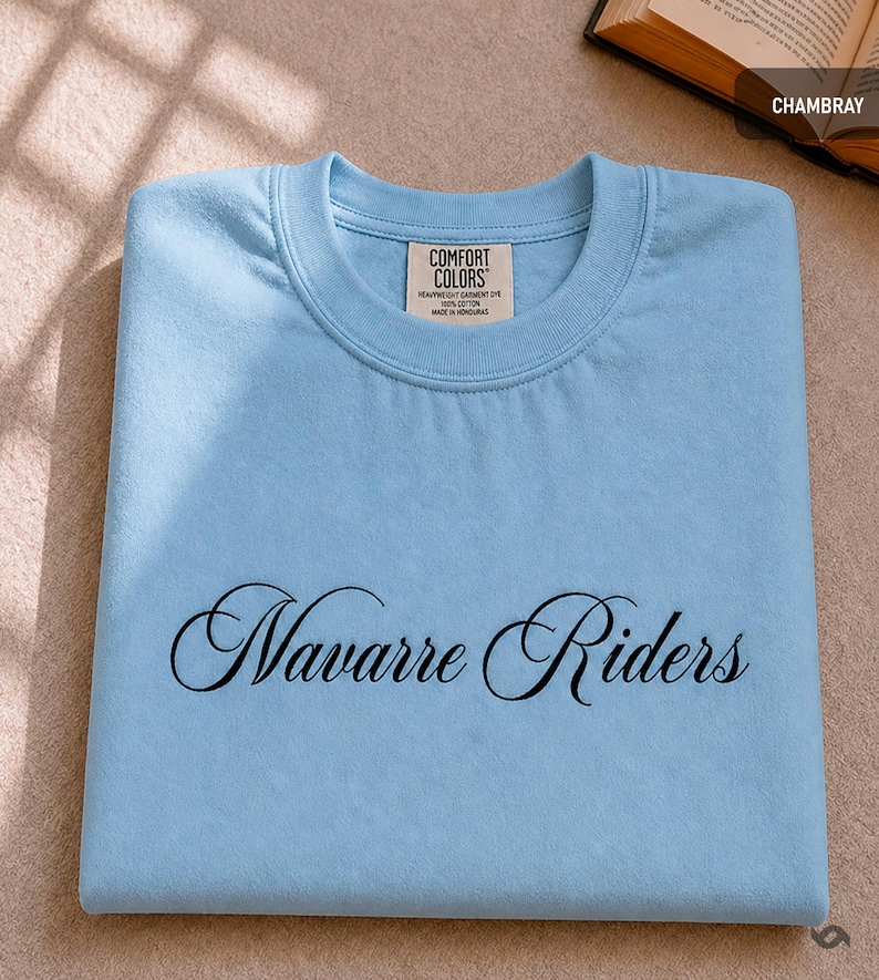 Puede incluir: Una camiseta azul claro con las palabras "Navarre Riders" en cursiva. La camiseta est&aacute; doblada cuidadosamente y tiene una etiqueta Comfort Colors. Est&aacute; hecha de un material de tinte para prendas pesadas.