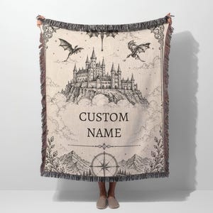 May include: A beige woven throw blanket featuring a detailed black and white illustration of a castle on a mountain, dragons, and the text "CUSTOM NAME". The blanket has a fringed edge and a compass design.