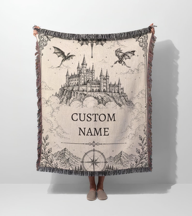 May include: A beige woven throw blanket featuring a detailed black and white illustration of a castle on a mountain, dragons, and the text "CUSTOM NAME". The blanket has a fringed edge and a compass design.
