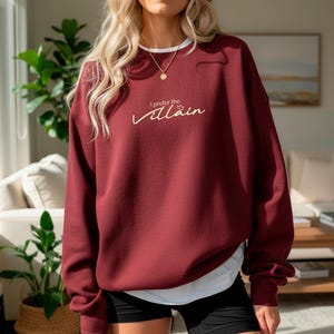 May include: A maroon sweatshirt with the text "I prefer the Villain" in a cursive font. The sweatshirt has a white collar and is paired with black shorts. A gold necklace is visible.