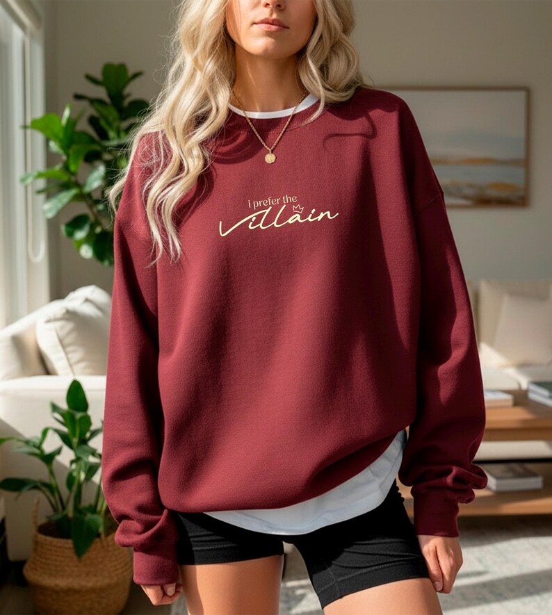 May include: A maroon sweatshirt with the text "I prefer the Villain" in a cursive font. The sweatshirt has a white collar and is paired with black shorts. A gold necklace is visible.