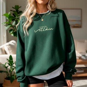 May include: A dark green sweatshirt with the text "I prefer the villain" in a cursive font. The sweatshirt has a white collar and is worn with black shorts. The overall style is casual and comfortable.