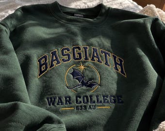 May include: A dark green crewneck sweatshirt featuring embroidered text that reads BASGIATH WAR COLLEGE 633 AU. The design includes a central yellow circular emblem with a star at the top and a dark blue dragon silhouette positioned in the middle.