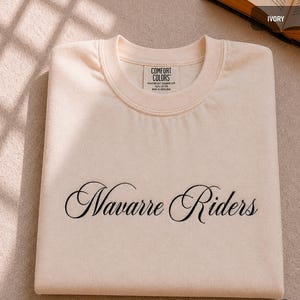 Puede incluir: Sudadera de color marfil con las palabras "Navarre Riders" bordadas en escritura negra. La sudadera tiene cuello redondo y una etiqueta que dice "Comfort Colors". La prenda est&aacute; doblada cuidadosamente.