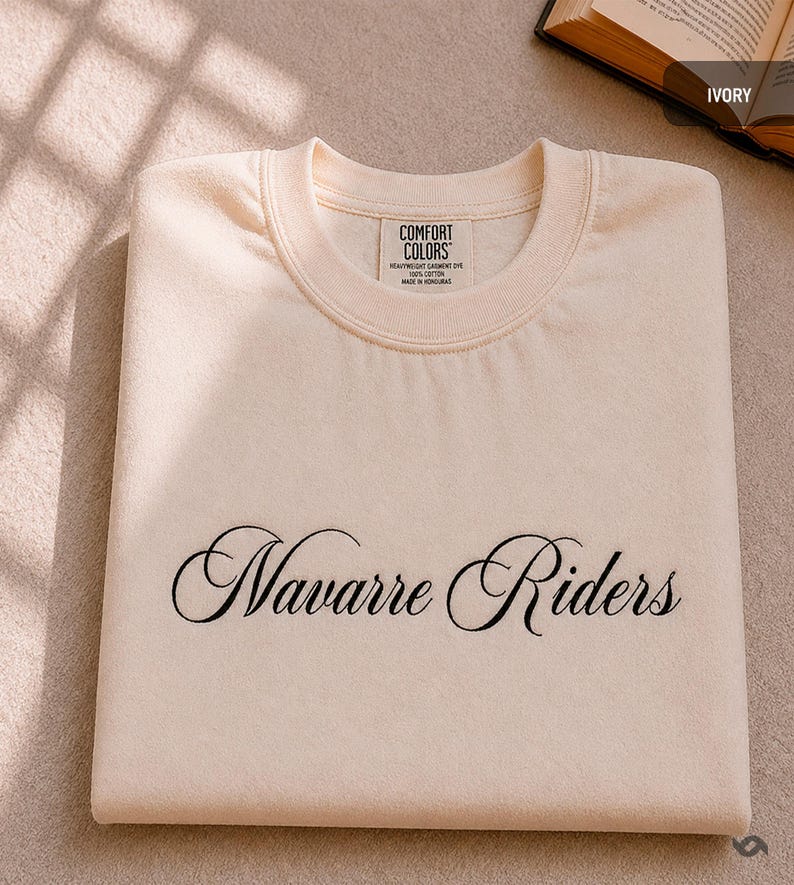 Puede incluir: Sudadera de color marfil con las palabras "Navarre Riders" bordadas en escritura negra. La sudadera tiene cuello redondo y una etiqueta que dice "Comfort Colors". La prenda est&aacute; doblada cuidadosamente.