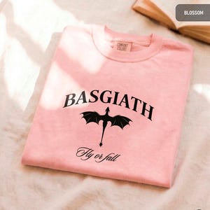 Peut inclure: Un sweat-shirt rose clair &agrave; col rond avec le mot "BASGIATH" en noir, en arc de cercle en haut, avec un dessin de dragon noir en dessous. L'expression "Fly or fall" est imprim&eacute;e en dessous du dragon. Le sweat-shirt est de Comfort Colors.