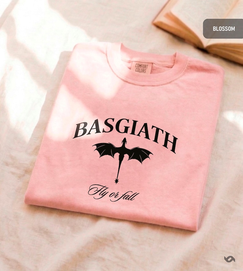 Peut inclure: Un sweat-shirt rose clair &agrave; col rond avec le mot "BASGIATH" en noir, en arc de cercle en haut, avec un dessin de dragon noir en dessous. L'expression "Fly or fall" est imprim&eacute;e en dessous du dragon. Le sweat-shirt est de Comfort Colors.