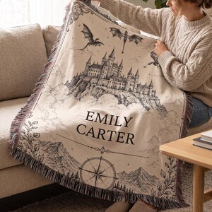 May include: Beige throw blanket with a black and white castle illustration, dragons, and the name "EMILY CARTER." The blanket has a fringed edge with shades of pink and purple. A compass rose design is at the bottom.
