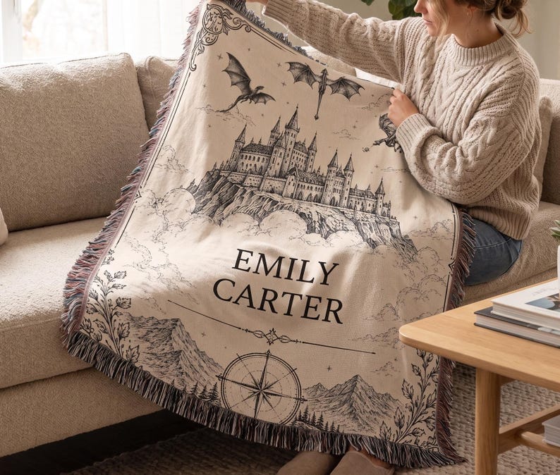 May include: Beige throw blanket with a black and white castle illustration, dragons, and the name "EMILY CARTER." The blanket has a fringed edge with shades of pink and purple. A compass rose design is at the bottom.