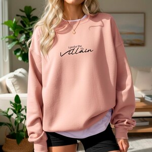 May include: A light pink sweatshirt with the text "I prefer the villain" in black script. The long-sleeved top is worn over a white shirt and black shorts. A gold necklace is visible.