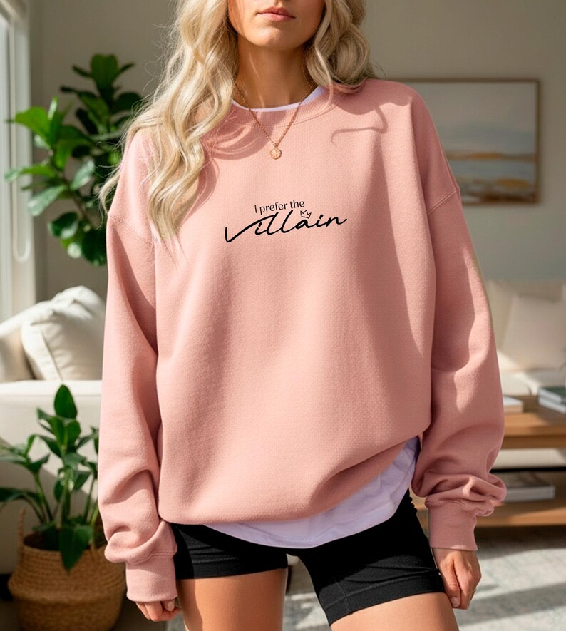 May include: A light pink sweatshirt with the text "I prefer the villain" in black script. The long-sleeved top is worn over a white shirt and black shorts. A gold necklace is visible.