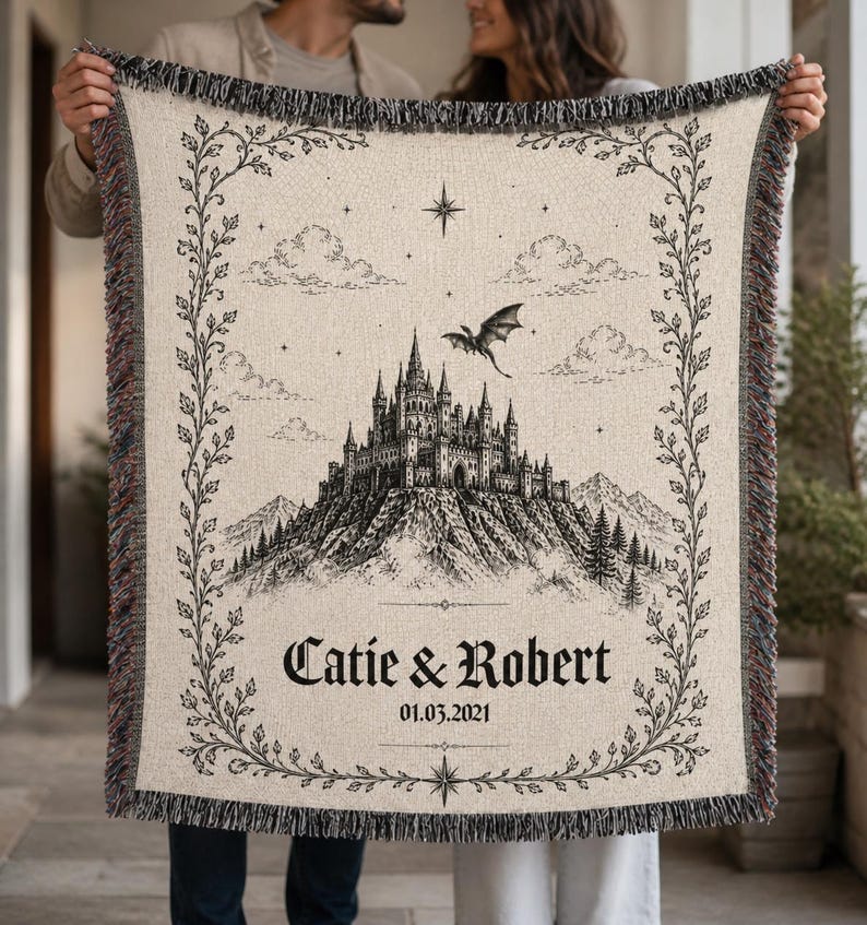 May include: Beige woven throw blanket featuring a black and white castle illustration with a dragon, clouds, and stars. The blanket has a decorative border and the names "Catie & Robert" with the date "01.03.2021". The edges have a fringed trim.