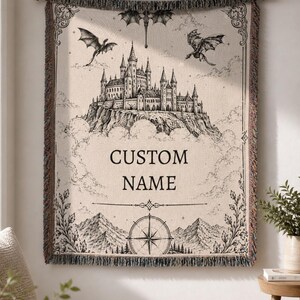 May include: A beige tapestry with a black and white castle illustration, dragons, and the text "CUSTOM NAME." The tapestry has a decorative border and is hanging on a wooden rod.