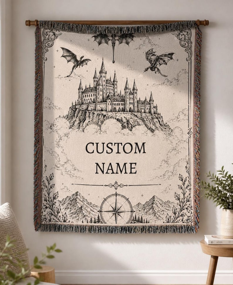 May include: A beige tapestry with a black and white castle illustration, dragons, and the text "CUSTOM NAME." The tapestry has a decorative border and is hanging on a wooden rod.