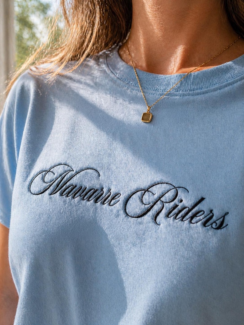 Puede incluir: Una camiseta azul claro con el texto bordado "Navarre Riders" en escritura negra. Se lleva un collar de oro con un colgante cuadrado. La camiseta es de material suave.