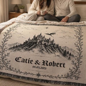 Op de afbeelding: Cr&egrave;mekleurige plaid met een zwart-wit illustratie van een kasteel op een berg, een draak en de namen "Catie & Robert" met de datum 01.03.2021. De plaid heeft een gefranjerde rand en een decoratieve rand.