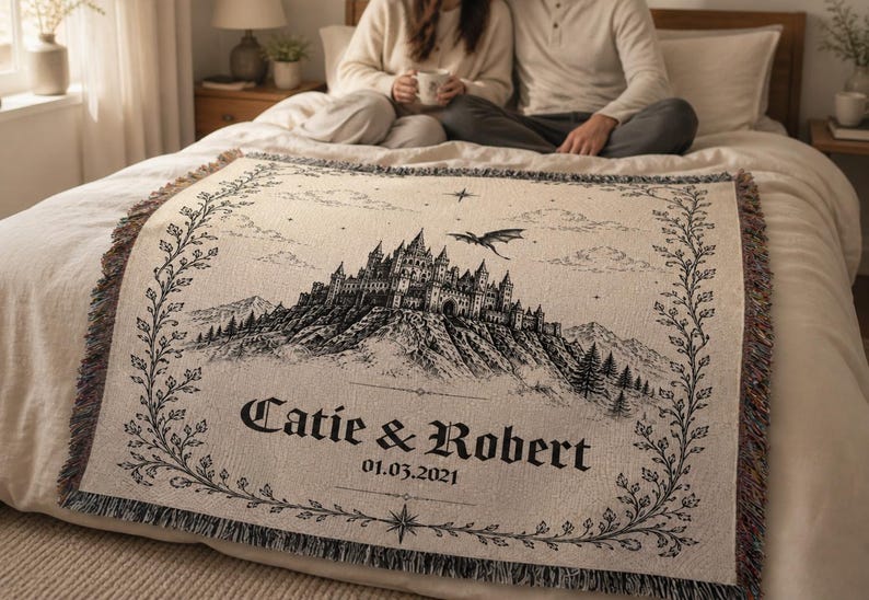 Op de afbeelding: Cr&egrave;mekleurige plaid met een zwart-wit illustratie van een kasteel op een berg, een draak en de namen "Catie & Robert" met de datum 01.03.2021. De plaid heeft een gefranjerde rand en een decoratieve rand.