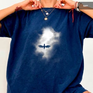 May include: Navy blue t-shirt featuring a dragon silhouette against a cloudy sky. The graphic is centered on the shirt. The shirt has short sleeves and a relaxed fit. The word "NAVY" is in the upper right corner.