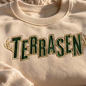 Puede incluir: Una sudadera de cuello redondo color crema con la palabra TERRASEN bordada en letras de bloque verde oscuro con contornos dorados. El texto está flanqueado por dos astas de ciervo bordadas en color dorado a los lados izquierdo y derecho.