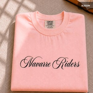 Puede incluir: Una sudadera rosa claro doblada con las palabras "Navarre Riders" en escritura negra en la parte delantera. La camisa tiene una etiqueta que dice "Comfort Colors".