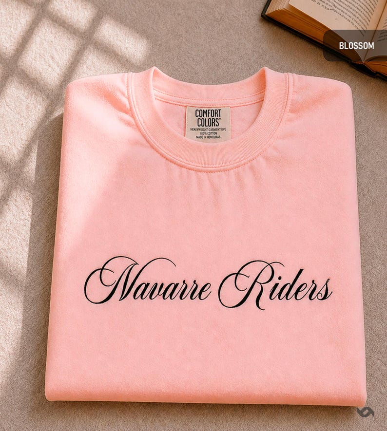 Puede incluir: Una sudadera rosa claro doblada con las palabras "Navarre Riders" en escritura negra en la parte delantera. La camisa tiene una etiqueta que dice "Comfort Colors".