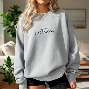 May include: A light gray sweatshirt with the text "I prefer the Villain" in black script. The sweatshirt has long sleeves and a crew neck. The model is wearing black shorts and a gold necklace.