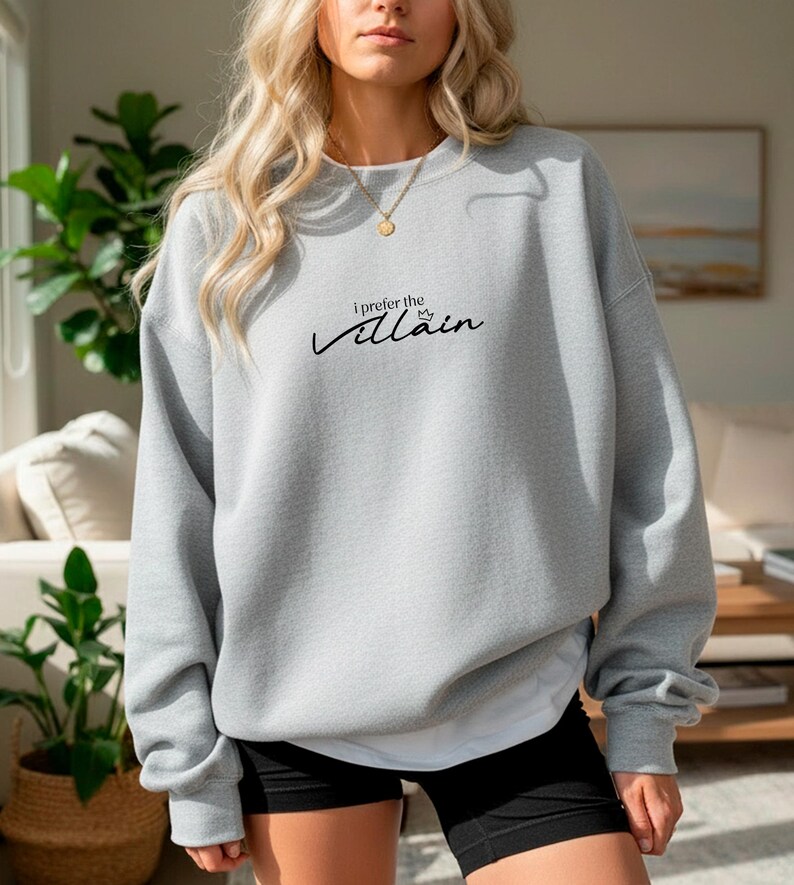 May include: A light gray sweatshirt with the text "I prefer the Villain" in black script. The sweatshirt has long sleeves and a crew neck. The model is wearing black shorts and a gold necklace.