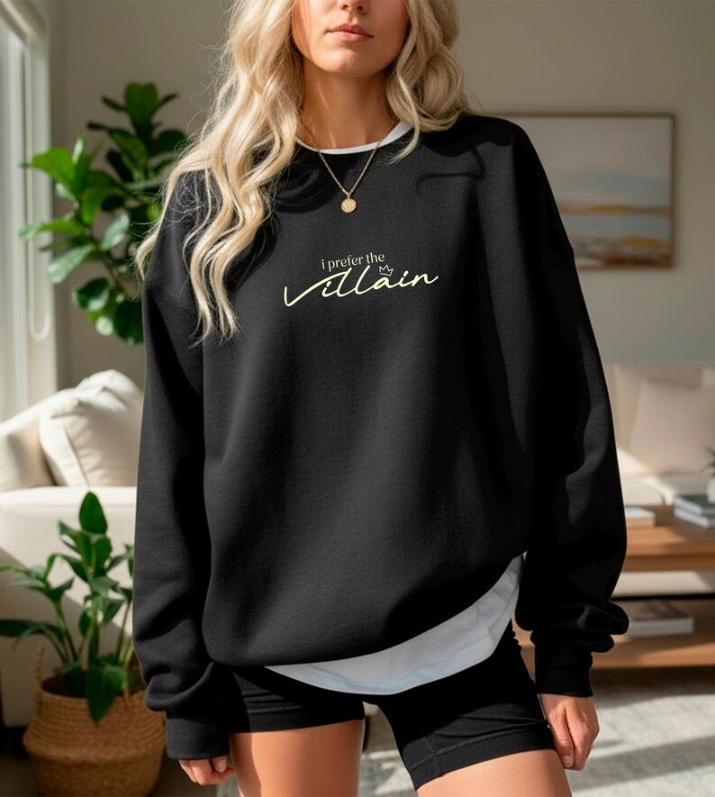 May include: Black sweatshirt with the text "I prefer the Villain" in a cursive font. The sweatshirt has a white crew neck and is layered over a white shirt. The model is wearing black shorts and a gold necklace.