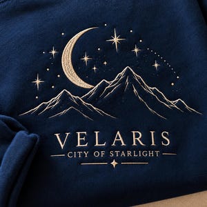 Könnte beinhalten: Ein dunkelblaues Sweatshirt mit goldener Stickerei, die eine Mondsichel, mehrere Sterne und ein Gebirge zeigt. Darunter befindet sich der in goldenen Großbuchstaben gestickte Text VELARIS CITY OF STARLIGHT mit einem kleinen Sternsymbol.