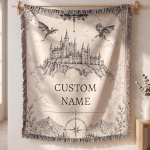 May include: Beige woven throw blanket with a black and white fantasy castle design, dragons, and the words "CUSTOM NAME". The blanket has a decorative border and fringe.