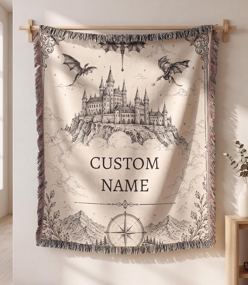May include: Beige woven throw blanket with a black and white fantasy castle design, dragons, and the words "CUSTOM NAME". The blanket has a decorative border and fringe.