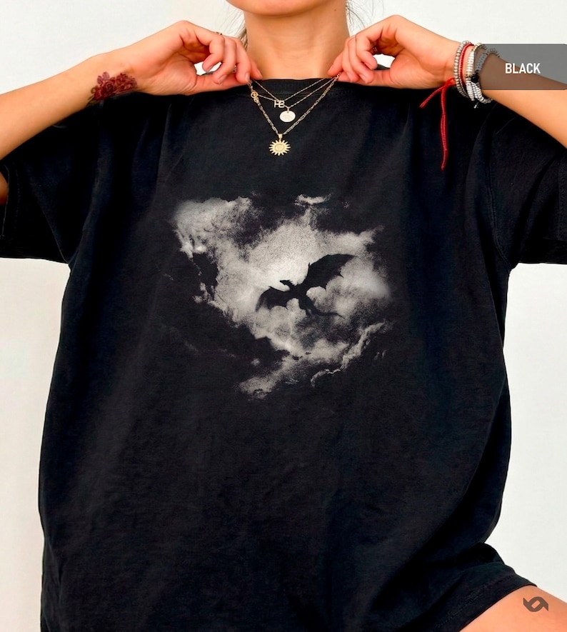 May include: Black t-shirt featuring a dragon silhouette against a cloudy background. The dragon is in flight, with its wings spread wide. The shirt is a dark color and has a classic crew neck design. The image is a graphic print.