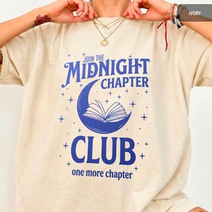Op de afbeelding: Ivoor kleurig t-shirt met de tekst "Join the Midnight Chapter Club" in blauw. Het ontwerp toont een halve maan met een open boek erin en sterren. De zin "one more chapter" is eronder gedrukt.