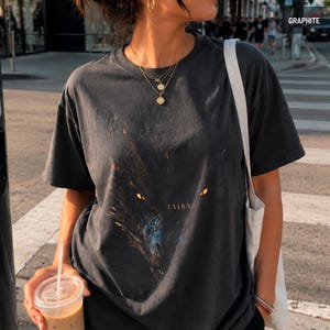 May include: Graphite-colored t-shirt featuring a wolf graphic with glowing yellow eyes and the word "TAIRN" in gold lettering. The shirt is a dark gray color and has a vintage, worn look.