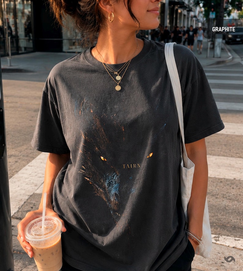 May include: Graphite-colored t-shirt featuring a wolf graphic with glowing yellow eyes and the word "TAIRN" in gold lettering. The shirt is a dark gray color and has a vintage, worn look.